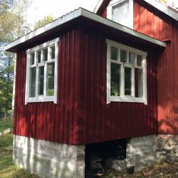 På billedet ser vi et lille rødmalede træhus med hvidt træværk omkring vinduerne. Huset er opført på en sokkel af grå betonsten og omgivet af grøn natur, som antyder at det er placeret i en have eller et skovområde. Farven på træbeskyttelsen er dyb og mættet kirsebær, hvilket fremhæver træets struktur og tilfører huset en klassisk og holdbar finish. 