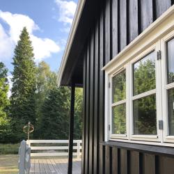 Du ser på en del af et hus omkranset af grønt landskab. Huset har en træfacade, som er malet i en dyb sort nuance, der står i kontrast til de hvide vinduesrammer. En veranda foran huset har hvide gelændere, hvilket komplementerer vinduernes stil. Ovenfor mødes en skyfri himmel, mens træer i forskellige grønne nuancer omgiver huset og skaber en rolig atmosfære. Billedet formidler den beskyttende effekt og det æstetiske udtryk, vores træmalingstilbud kan bidrage med til dit hjem.