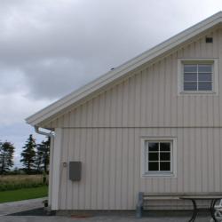På billedet ser du en ydersiden af et hus, der står under en skyet himmel. Huset har lodrette træbeklædninger, og et simpelt, skråt tag. Der er ét synligt vindue med hvide rammer og en lys persienne bagved. En mindre bænk er placeret under vinduet. Farven på husets facade forekommer at være en slags lys beige eller lysegul, en farve der vil falde godt ind i vores træbeskyttelsesmaling-kollektion. Uden yderligere farveoplysninger kan det egentlige nuance ikke specificeres. Husket, malingsspandene på vores webshop indeholder 5 liter maling hver.