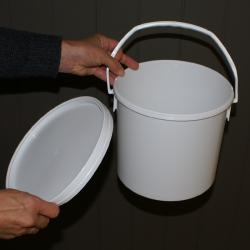 En person holder låget til en hvid plastikspand i den ene hånd. Spanden har et hvidt håndtag og står på en mørkegrå baggrund.