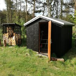 Dette billede viser to små skure i en have, det ene med åben dør, det andet delvist fyldt med stablet brænde. Skuret i forgrunden ser ud til at være brugt til opbevaring og har en rød genstand inde i sig. Rundt om skurene er grønt græs, og bag dem ses træer. Det er en solrig dag.